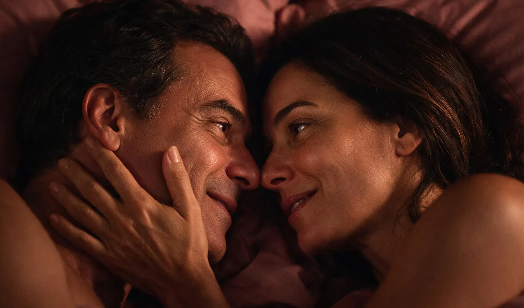 O mesmo casal deitado na cama em luz quente, frente a frente, muito próximos, ela com a mão no rosto dele