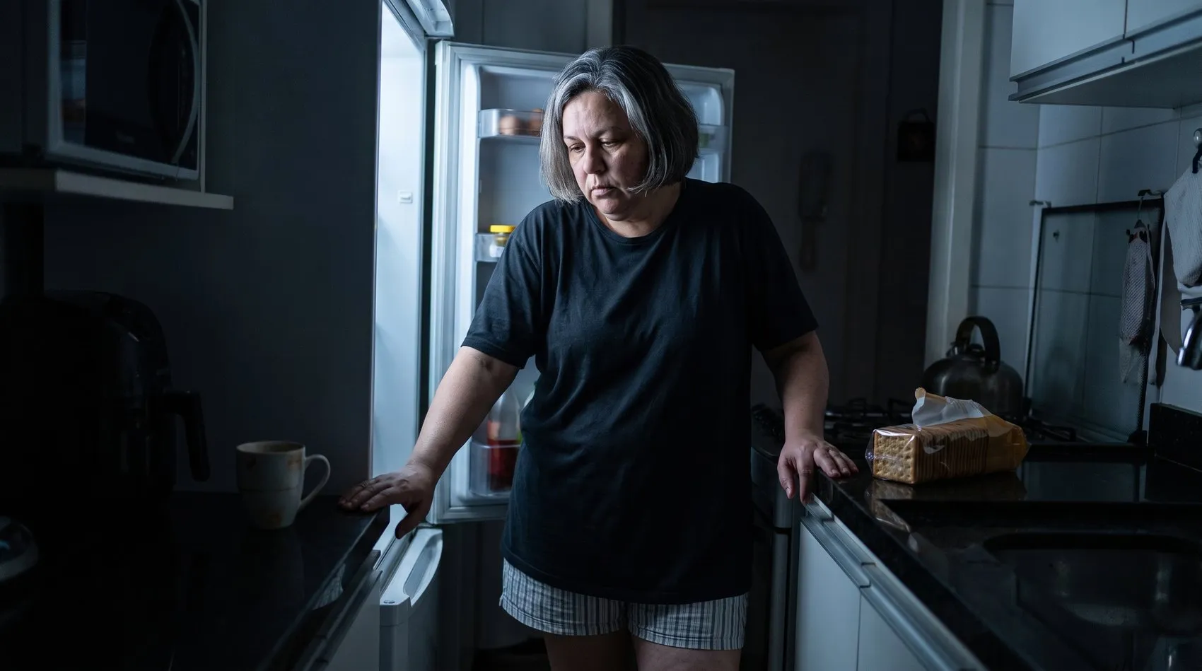 A mesma mulher de madrugada na cozinha escura, de pijama, parada diante da geladeira aberta com a mão na porta, olhando para dentro sem escolher nada