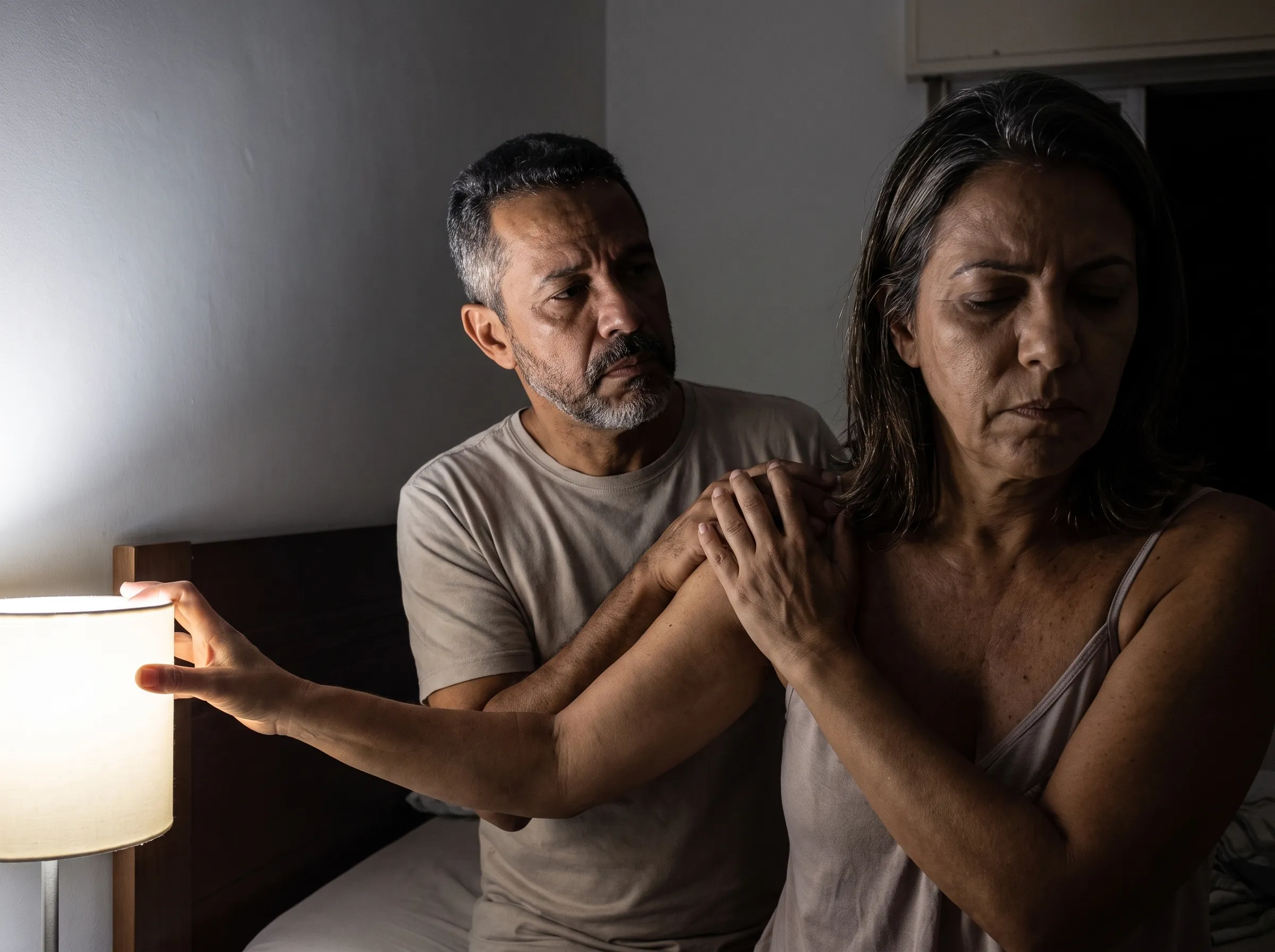 Quarto à noite: o marido põe a mão no ombro dela e ela, de rosto fechado, tira a mão dele do ombro enquanto estende o braço para apagar o abajur