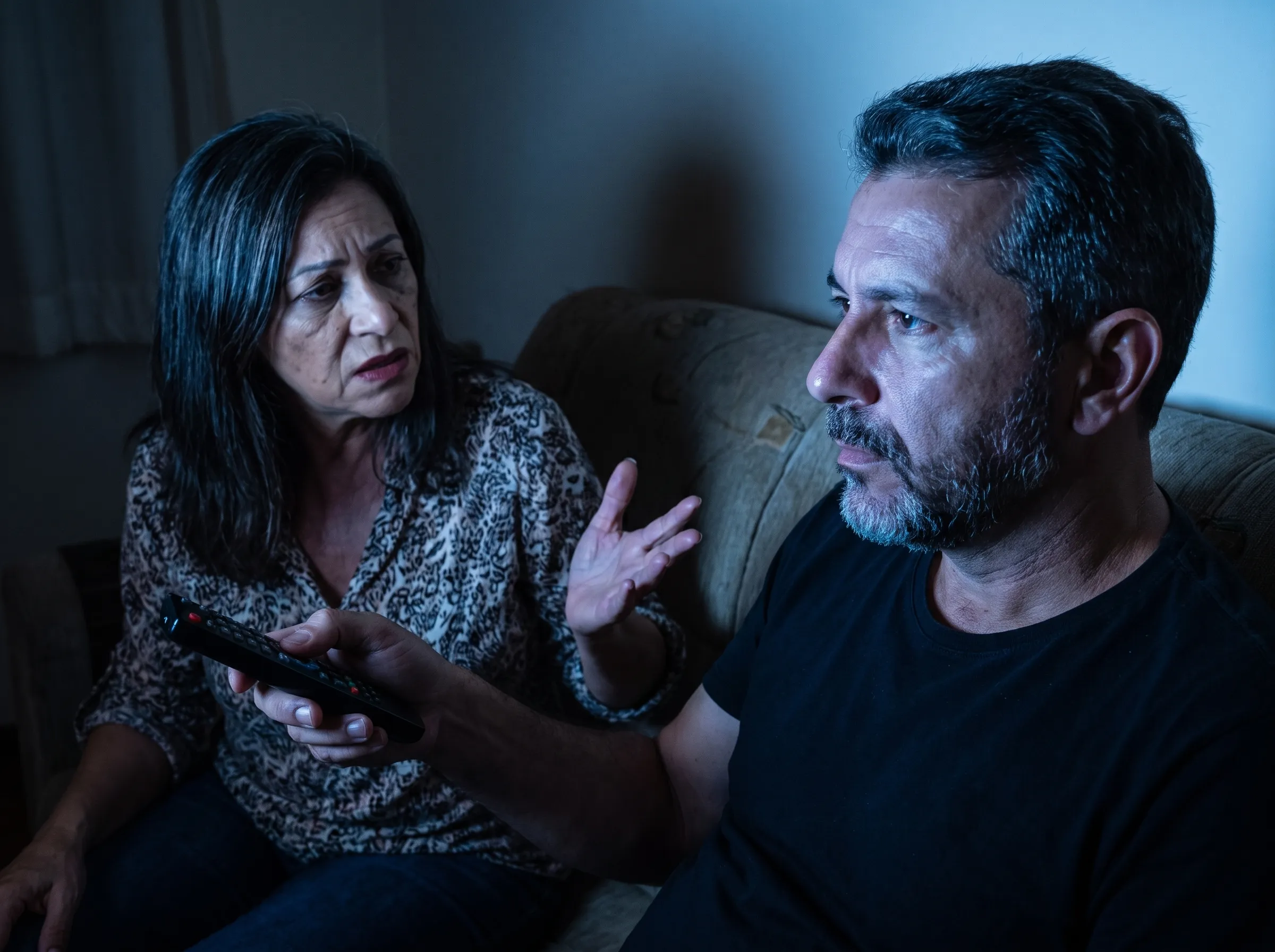Sala à noite: o marido no sofá, de cabeça virada para a televisão fora do quadro, com o controle na mão; ela, na outra ponta do sofá, fala com ele de mão erguida e ele não olha